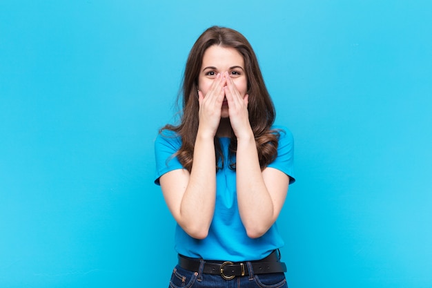 Jeune jolie femme à la bouche heureuse, joyeuse, chanceuse et surprise couvrant les deux mains sur le mur bleu