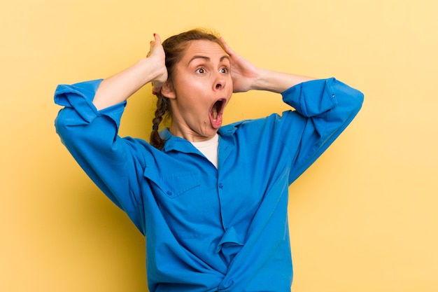 Jeune jolie femme à la bouche ouverte qui a l'air horrifiée et choquée à cause d'une terrible erreur levant les mains à la tête