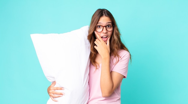 Jeune jolie femme avec la bouche et les yeux grands ouverts et la main sur le menton portant un pyjama et tenant un oreiller