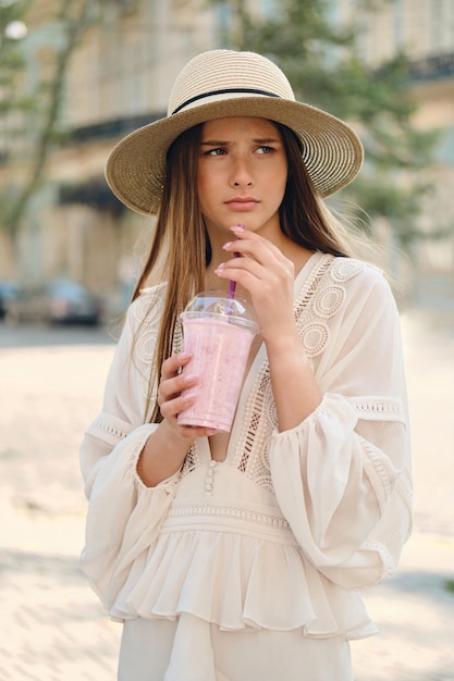 Jeune jolie femme bouleversée en robe blanche et chapeau tenant un smoothie pour aller dans la main regardant tristement de côté tout en se tenant seul dans la rue de la ville