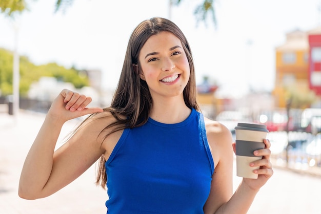 Jeune jolie femme brésilienne tenant un café à emporter à l'extérieur fière et satisfaite