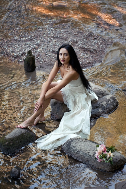 Jeune jolie femme brune en robe de mariée blanche assis près de la rivière de la forêt et souriant