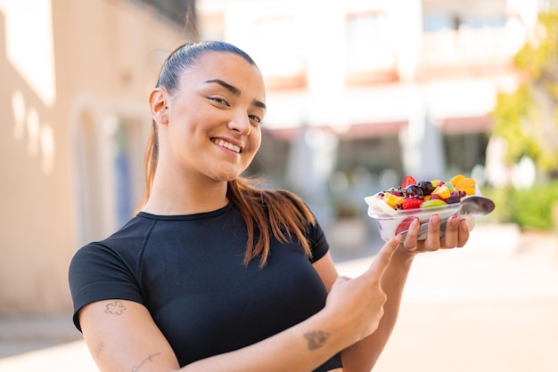 Jeune jolie femme brune tenant un bol de fruits à l'extérieur et le pointant