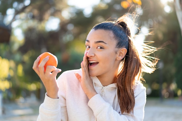 Jeune jolie femme brune tenant une orange à l'extérieur en chuchotant quelque chose