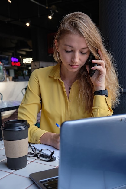 Jeune jolie femme caucasienne assise avec un ordinateur portable au café appelant par smartphone