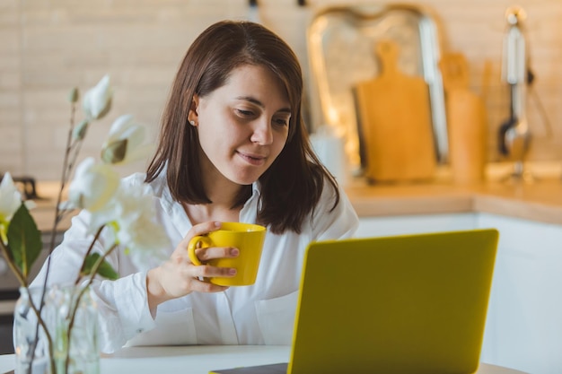 Jeune jolie femme caucasienne parlant au téléphone assise devant un ordinateur portable dans la cuisine