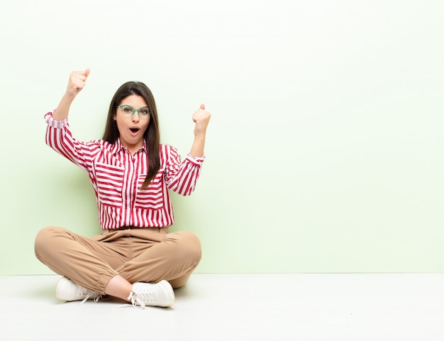 Jeune jolie femme célébrant un succès incroyable comme une gagnante, l'air excitée et heureuse de dire ça! assis par terre