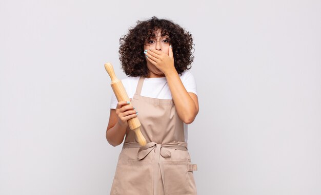 Jeune jolie femme chef couvrant la bouche avec les mains avec une expression choquée et surprise, en gardant un secret ou en disant oups