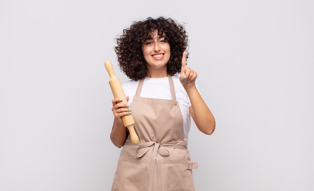 Jeune jolie femme chef souriant et à la sympathique, montrant le numéro un ou d'abord avec la main en avant, compte à rebours