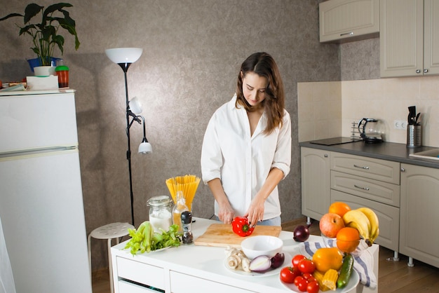 Une jeune jolie femme en chemise blanche dans la cuisine coupe des légumes poivrons rouges maïs oignons bleus Végétariens aliments sains