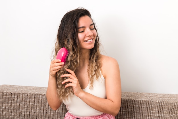 Jeune jolie femme avec des cheveux bouclés les brosser tout en souriant sur le canapé à la maison