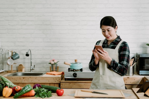 Jeune jolie femme chinoise asiatique utilisant un smartphone debout à la table de comptoir pleine de fruits et légumes dans l'intérieur de la cuisine en bois. Compter les calories avec l'application pour téléphone portable. Concept de soins de santé alimentaire