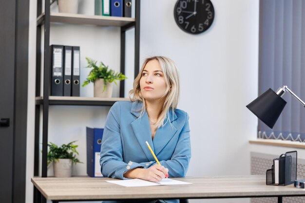 Jeune jolie femme en costume assis au bureau au bureau tenant un crayon et prenant des notes dans un cahier