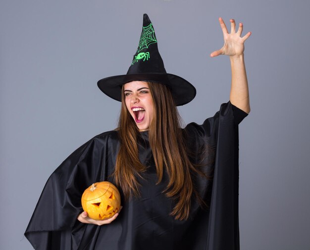 Jeune jolie femme en costume de sorcière avec chapeau noir tenant une citrouille sur fond bleu en studio