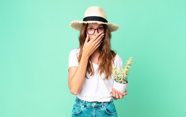 Jeune jolie femme couvrant la bouche avec les mains avec un choqué avec un chapeau de paille et tenant un cactus