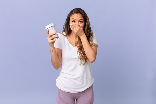 Jeune jolie femme couvrant la bouche avec les mains avec un choqué et tenant un café