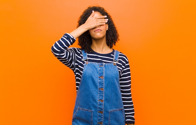 Jeune jolie femme couvrant les yeux avec une main se sentant effrayé ou anxieux, se demandant ou attendant aveuglément une surprise sur le mur orange