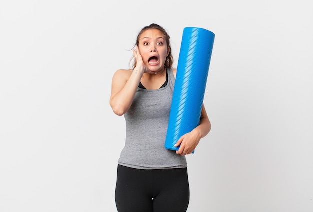 Jeune jolie femme criant avec les mains en l'air et tenant un tapis de yoga