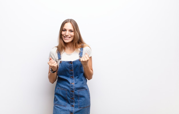 Jeune jolie femme criant triomphalement, riant et se sentant heureux et excité tout en célébrant le succès contre le mur blanc