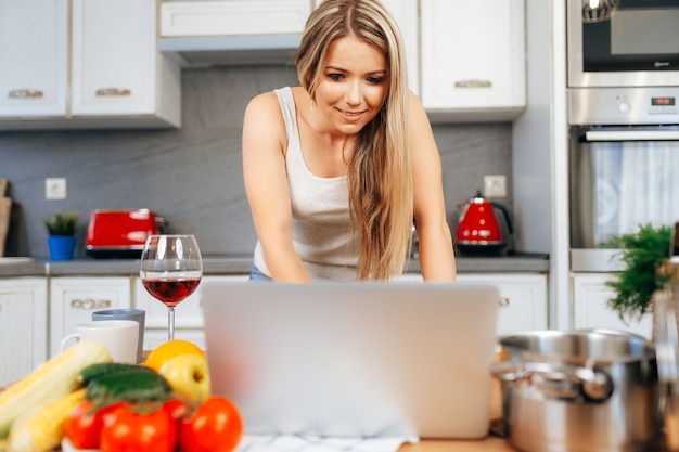 Jeune jolie femme cuisine quelque chose dans sa cuisine avec ordinateur portable