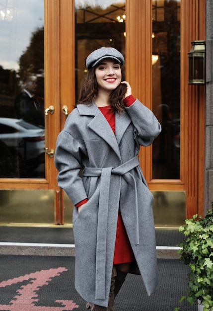 Jeune jolie femme élégante vêtue d'une robe rouge, d'un manteau gris et d'un chapeau posant dans les rues de la ville