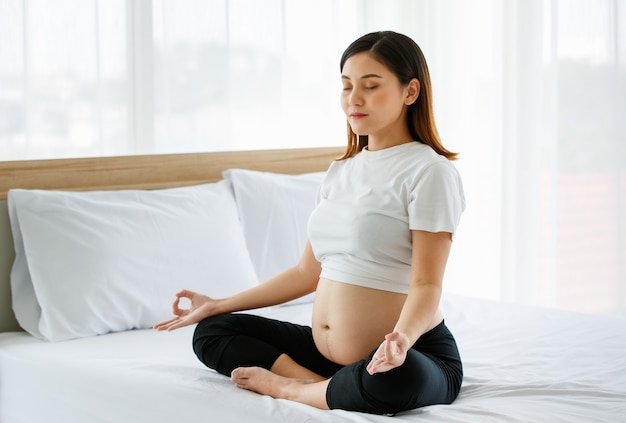 Jeune jolie femme enceinte asiatique assise sur un lit faisant de l'exercice délicieusement par le yoga. Elle sourit et regarde une caméra. Concept de mère heureuse en bonne santé.