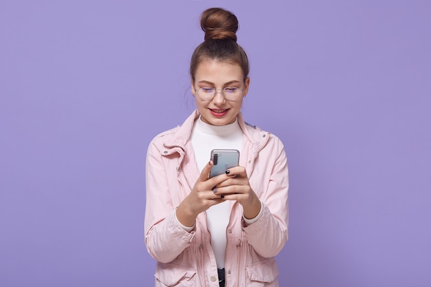 Jeune jolie femme européenne isolée sur fond lilas debout en veste rose pâle, textos sur téléphone portable avec compagnon, montrant un sourire amical heureux.