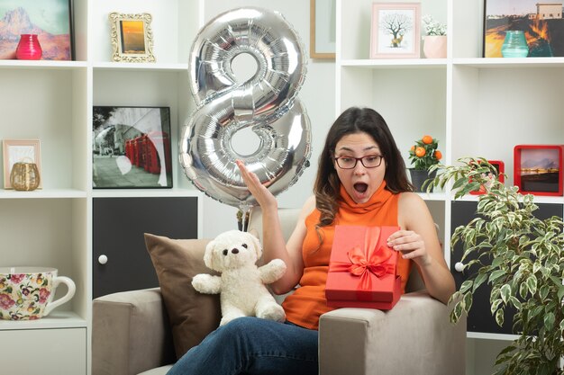 Jeune Jolie Femme Excitée Dans Des Verres S'ouvrant Et Regardant Une Boîte-cadeau Assise Sur Un Fauteuil Dans Le Salon Le Jour De La Journée Internationale De La Femme En Mars