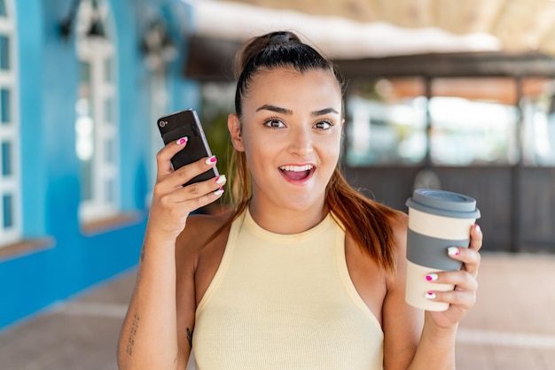 Jeune jolie femme à l'extérieur utilisant un téléphone portable et tenant un café avec une expression heureuse