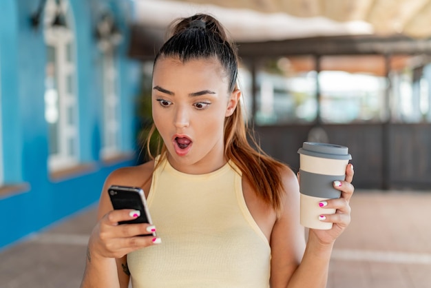 Jeune jolie femme à l'extérieur utilisant un téléphone portable et tenant un café avec une expression surprise