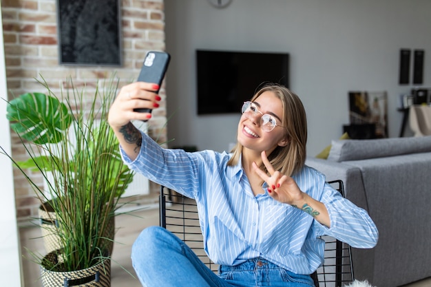 Jeune jolie femme avec un geste de paix assis dans une chaise prenant selfie près de la fenêtre à la maison.