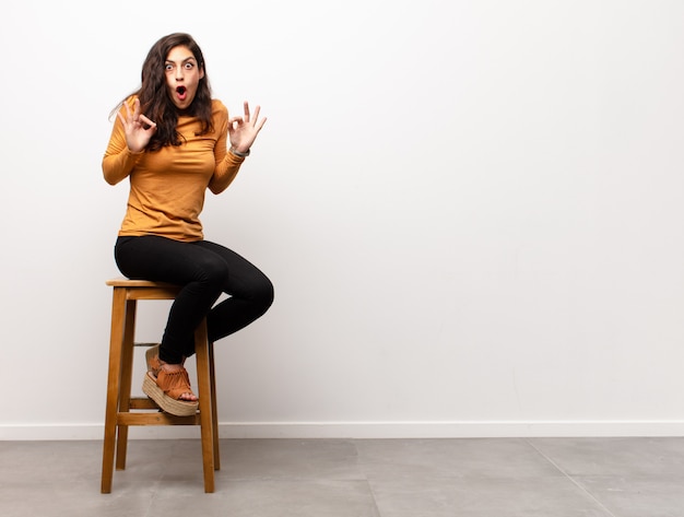 Jeune jolie femme heureuse et excitée, surprise et étonnée couvrant la bouche avec les mains, riant avec une expression mignonne assise dans une pièce