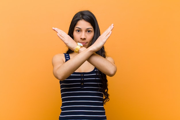 Jeune jolie femme hispanique à l'air agacée et malade de votre attitude, en dit assez! les mains croisées sur le devant, vous disant de vous arrêter contre le mur brun