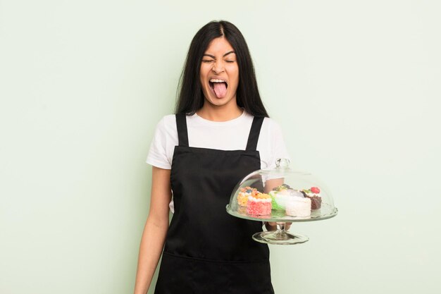 Jeune jolie femme hispanique avec une attitude joyeuse et rebelle plaisantant et tirant la langue chef avec concept de gâteaux
