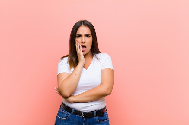 Jeune jolie femme hispanique bouche bée de choc et d'incrédulité, la main sur la joue et le bras croisés, se sentant stupéfaite et étonnée contre le mur rose