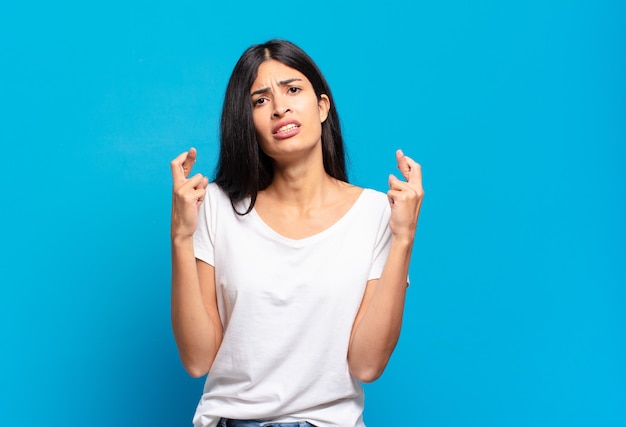 Jeune jolie femme hispanique croisant les doigts avec anxiété et espérant avoir de la chance avec un regard inquiet