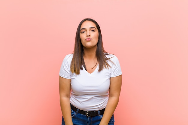 Jeune jolie femme hispanique pressant les lèvres avec une expression mignonne, amusante, heureuse et charmante, envoyant un baiser contre le mur rose