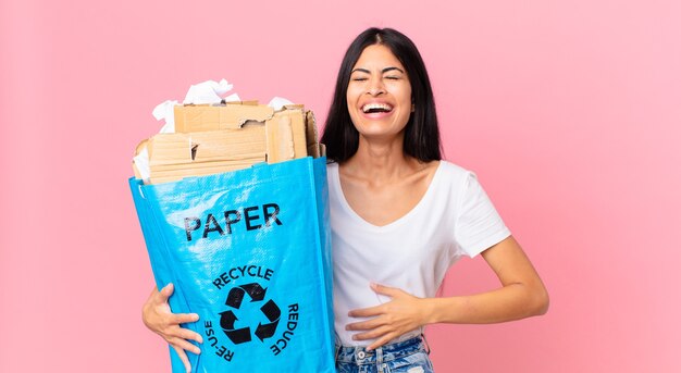 Jeune jolie femme hispanique riant aux éclats d'une blague hilarante et tenant un sac en papier à recycler