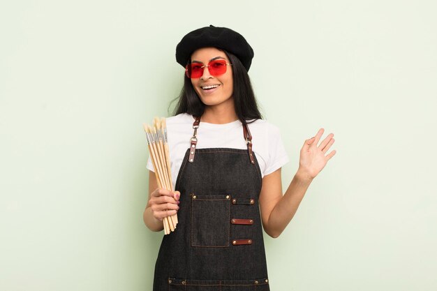 Jeune jolie femme hispanique souriant joyeusement, agitant la main, vous accueillant et vous saluant. concept d'étudiant en art