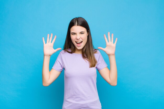 Jeune jolie femme hurlant de panique ou de colère, choquée, terrifiée ou furieuse, les mains à côté de la tête contre le mur bleu