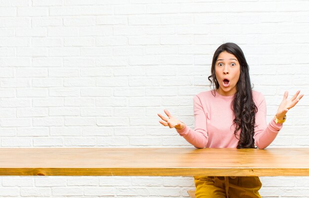 Jeune jolie femme latine bouche bée et émerveillée, choquée et étonnée d'une incroyable surprise assise devant une table