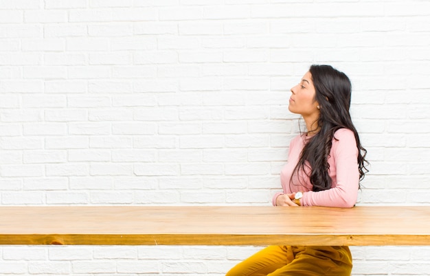 Jeune jolie femme latine se sentant confuse ou pleine ou des doutes et des questions, se demandant, avec les mains sur les hanches, vue arrière assis devant une table