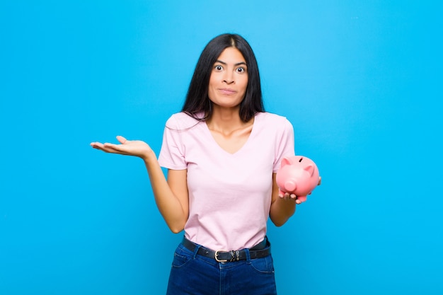 Jeune jolie femme latine se sentant perplexe et confuse, doutant, pondérant ou choisissant différentes options avec une drôle d'expression contre un mur plat