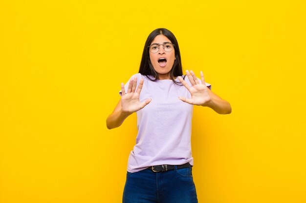 Jeune jolie femme latine se sentant stupéfaite et effrayée, craignant quelque chose d'effrayant, les mains ouvertes devant lui disant de rester à l'écart du mur plat