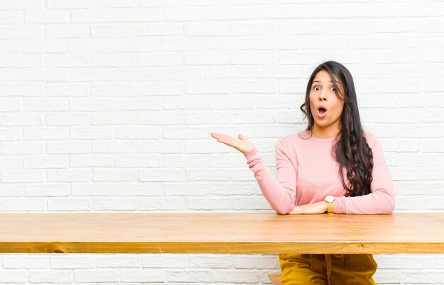 Jeune jolie femme latine semblant surprise et choquée, avec la mâchoire tombée, tenant un objet avec une main ouverte sur le côté, assise devant une table