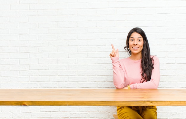 Jeune jolie femme latine souriante gaie et joyeuse, pointant vers le haut avec une main pour copier l'espace assis devant une table