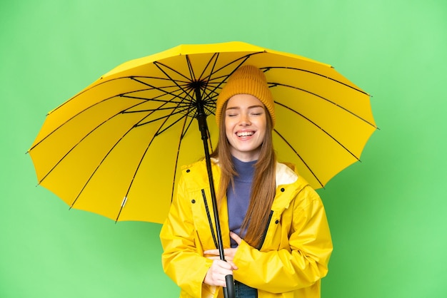 Jeune jolie femme avec manteau imperméable et parapluie sur fond isolé chroma key souriant beaucoup