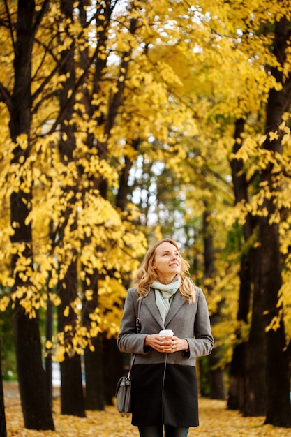 Jeune jolie femme marchant dans le parc en automne avec une tasse de café