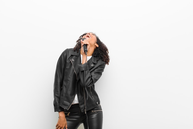 Jeune jolie femme noire avec un microphone portant une veste en cuir contre le mur blanc