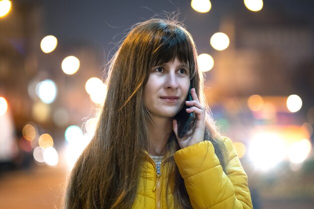 Jeune jolie femme parlant sur son téléphone portable debout dans la rue de la ville la nuit à l'extérieur.